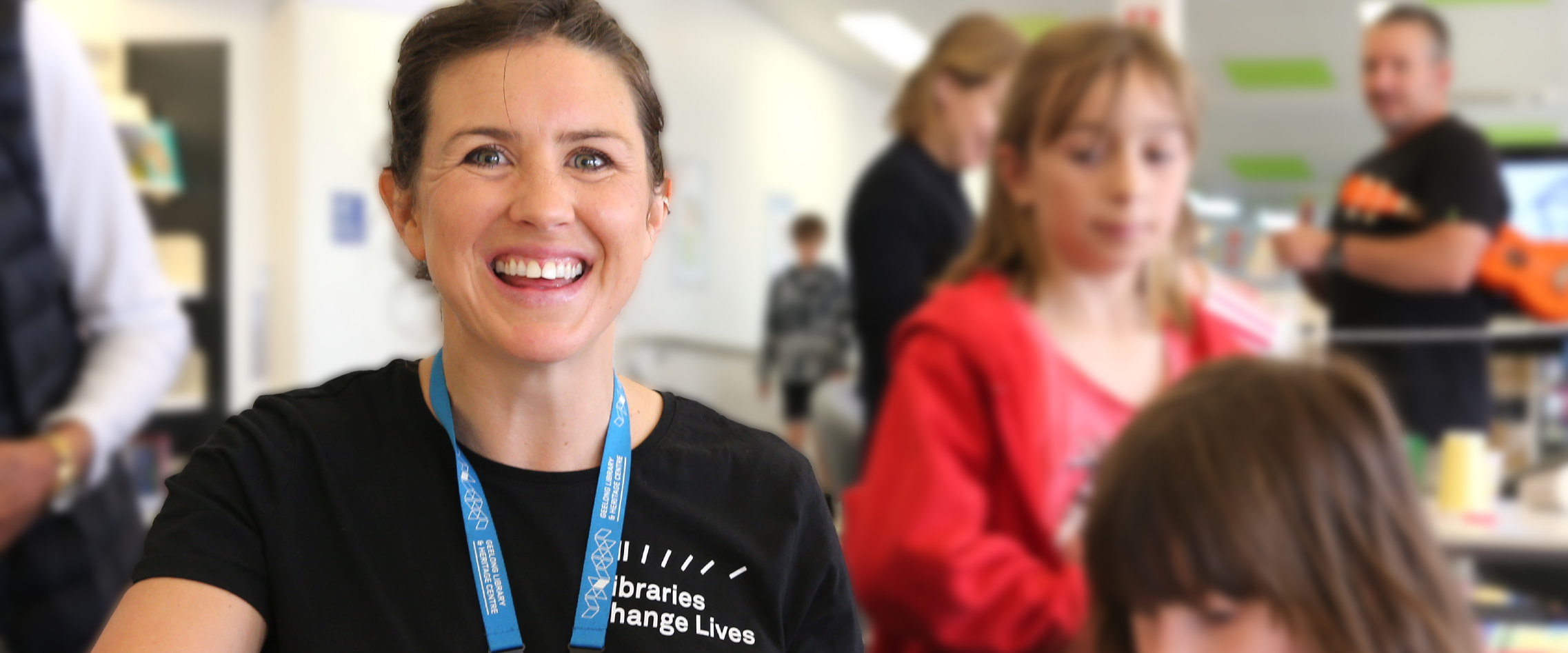 A young woman in library smiles at the camera, there are children around her.