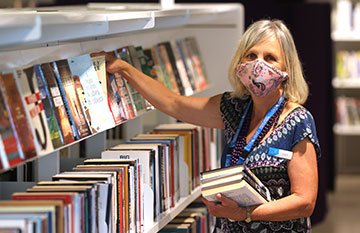 Image description: A blond woman wearing a face mask looks at the camera while placing books on the shelf.