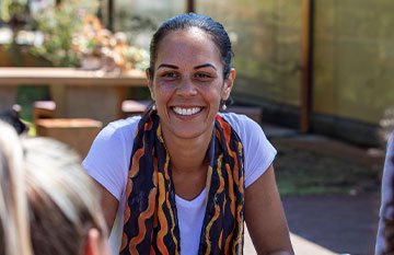 Image description: A woman with dark hair and eyes smiles sitting outdoors. She is wearing a scarf and white tshirt.