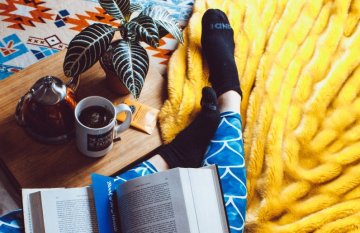 birds-eye view of a woman's legs with open book. She is sitting on a yellow rug with a cup of tea nearby